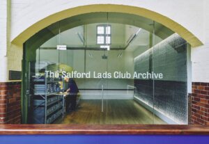 A wide shot of an archive space filmed through an archway and glass wall. Etched on to the glass is white text: 'The Salford Lads Club Archive'.