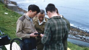 Four men stand in a tight circle holding something. They're stood on a cliff edge by the sea.