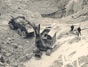 A black and white photograph of a steam shoveller, a truck and two people in a working pit.