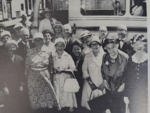 A black and white photograph showing at least a dozen people (predominantly elderly) stood smiling in front of a bus.