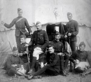 A black and white photograph of several uniformed soldiers posing for the camera outside of a white tent. Two of the men have small dogs on their laps.