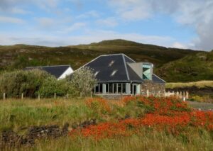 A wide shot of a small building surrounded by bucolic mountains, grassland and bright red flowers.